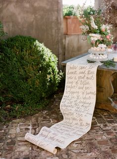 a table with a sign on it sitting in front of some bushes and flowers next to a potted plant