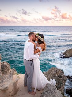 a bride and groom kissing on the rocks by the ocean at sunset in front of some cliffs