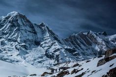 two people walking up the side of a snow covered mountain in front of a cloudy sky