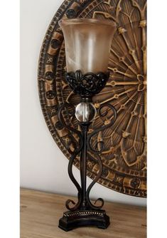 a candle holder with a glass on it in front of a wall hanging plaque that has an ornate design