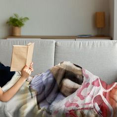 a woman laying on top of a couch under a blanket next to a pillow holding a book