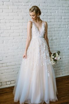 a woman standing in front of a white brick wall wearing a wedding dress with flowers on it