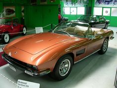 an orange sports car is on display in a green room with other classic cars behind it