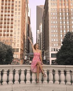 a woman in a pink dress sitting on the edge of a fountain with her hand up