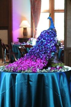 a peacock statue sitting on top of a table covered in purple flowers and greenery