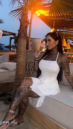 a woman in white dress sitting next to a palm tree and drinking from a cup