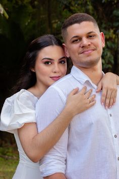 a young man and woman are posing for a photo in front of some trees with their arms around each other