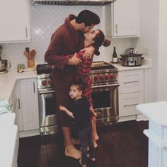 a man and woman kissing in the kitchen with their child on his lap, standing next to an oven