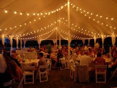 a group of people sitting at tables under a white tent with lights strung over it