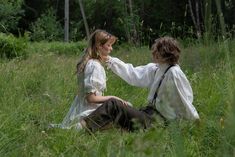 two people sitting on the ground in tall grass, one is touching the other's hand