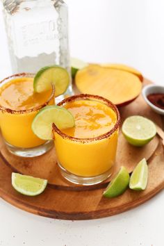 two glasses filled with orange juice and limes on a wooden platter next to condiments