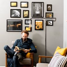 a man sitting on a chair reading a book in front of a wall full of pictures