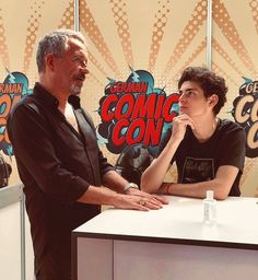 two men standing next to each other in front of a comic con booth with microphones