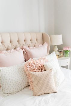 a white bed topped with lots of pillows next to a pink headboard and lamp