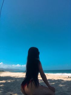 a woman sitting on top of a sandy beach next to the ocean under a blue sky