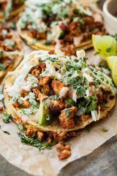 some tacos with meat and vegetables on top of paper next to lemon wedges