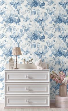 a white dresser sitting next to a wall with blue flowers on it and a lamp