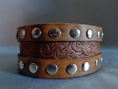 Two leather bands are riveted outside of a Victorian scroll pattern on this medium brown leather band. Adjustable snap closure. Western Style Brown Cuff Bracelet For Festivals, Artisan Brown Concho Cuff Bracelet, Brown Leather Bracelet With Concho, Artisan Brown Cuff Bracelet With Concho, Brown Leather Concho Bracelet, Western Brown Hand-tooled Leather Bracelet, Adjustable Brown Leather Bracelet With Custom Hardware, Rustic Brown Cuff Bracelet With Concho, Adjustable Brown Western Cuff Bracelet