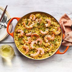 an orange pan filled with shrimp and rice on top of a white table next to silverware