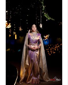 a woman in a purple and gold sari standing under a tree with lights behind her