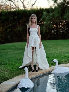 a woman in a white dress standing next to two swans on the edge of a pool