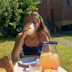 a woman sitting at an outdoor table drinking from a pitcher and glasses filled with lemonade