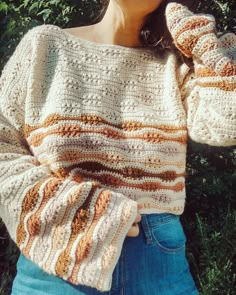 a woman wearing a white sweater and blue jeans standing in front of some bushes with her hand on her shoulder