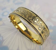 "The Binder Bros. Inc. (est. 1920 NY, NY) made this antique 10kt gold filled bangle with a lovely chased and engraved floral leaf vine design. Both sides of the form are covered with lots of detail and a patterned background. A lot of the original black enamel is present with some loss. This piece looks beautiful on its own, but would also be a fun stacking bangle. It has a working push button clasp. The original safety chain is missing so I have included a free 17 1/2 inch 12kt gold filled chai Victorian Engraved Bangle For Gift, Antique Engraved Gold Bracelet For Wedding, Victorian Etched Yellow Gold Bangle, Antique Etched Bangle For Wedding, Ornate Engraved Yellow Gold Bangle, Antique Etched Wedding Bangle, Victorian Style Yellow Gold Bangle Gift, Heirloom Filigree Bangle Gift, Heirloom Filigree Bangle As A Gift