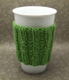 a green knitted cup sleeve sitting on top of a white coffee cup next to a wall