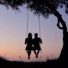 two people sitting on swings under a tree at sunset or dawn with the sun setting behind them