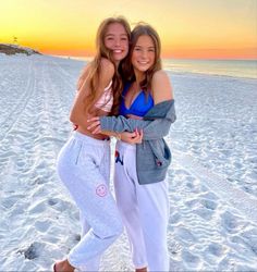 two girls are hugging on the beach at sunset