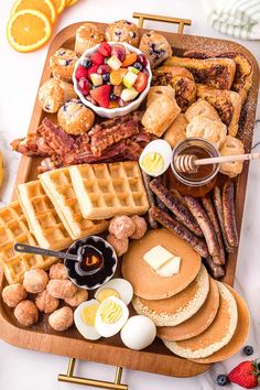 an assortment of breakfast foods including waffles, bacon, eggs and fruit on a wooden platter