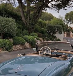 an old blue convertible car parked on the side of a road next to trees and bushes