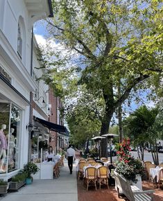 people are walking down the sidewalk in front of shops on a sunny day with lots of greenery