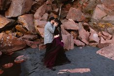 a man standing next to a woman on top of a rock covered beach with rocks in the background