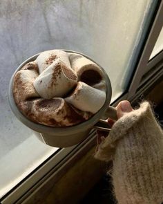 a person holding a cup filled with hot chocolate and marshmallows on top of a window sill