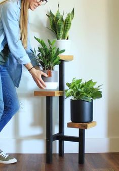 a woman standing next to two plants on top of a wooden table in front of a white wall