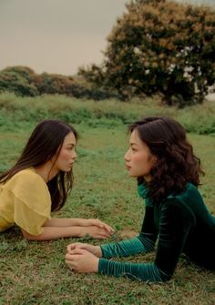 two young women laying on the ground talking to each other in an open field with trees and grass behind them