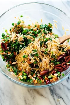 a glass bowl filled with pasta salad on top of a white marble countertop next to a wooden spoon