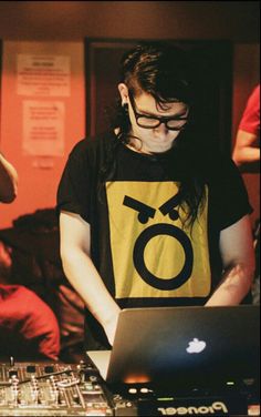 a man in glasses is working on his laptop at a dj's desk with other people behind him