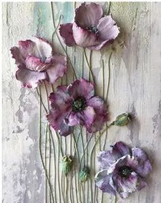 some purple flowers are on a white table and one is hanging off the wall with green stems