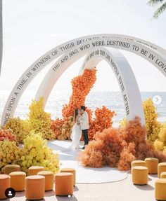 a bride and groom standing in front of an arch with orange flowers on it near the ocean