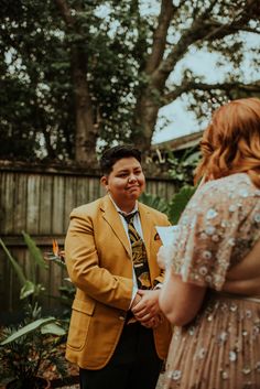 a man standing next to a woman in a dress and yellow blazer talking to each other