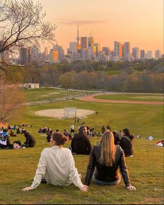 two people sitting on the grass in front of a large cityscape at sunset