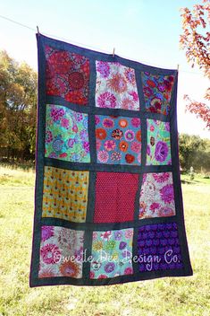 a patchwork quilt hanging on a clothesline in the grass with trees in the background