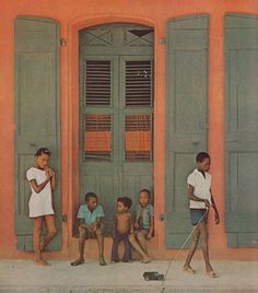 children are standing in front of an orange building with shutters and green doors,