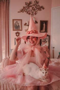 a woman in a pink dress and hat with a white cat on her lap sitting on a bed