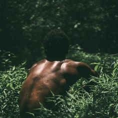 a shirtless man standing in the middle of tall grass and trees, looking down at his back