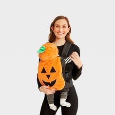 a woman is holding a baby in a pumpkin costume while wearing a black jacket and leggings