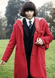 a woman in a red coat is standing on the grass with her hands behind her back